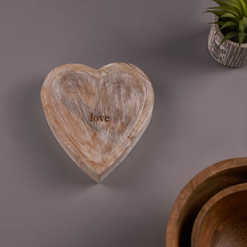 A heart-shaped wooden decor with a rustic finish, placed on a flat surface with a plant in the background.