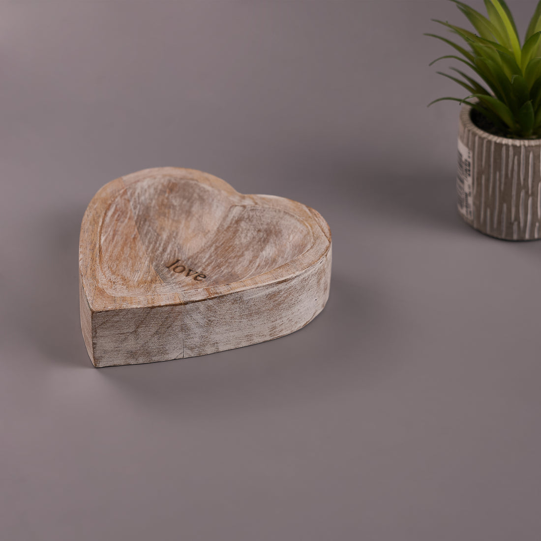 A heart-shaped wooden decor with a rustic finish, placed on a flat surface with a plant in the background.