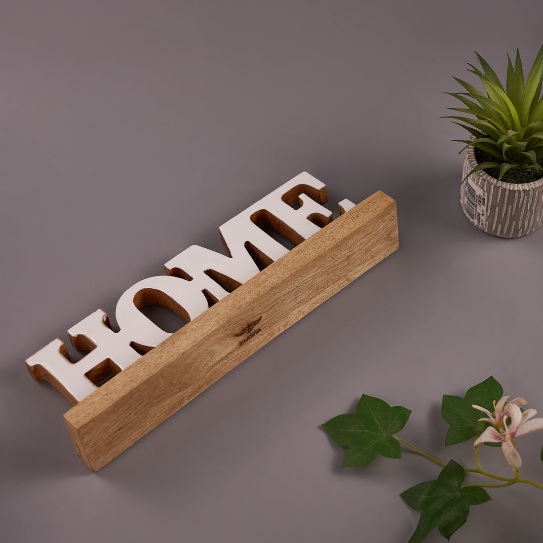 A wooden 'HOME' word sign with a natural finish, displayed on a gray surface with a potted plant and decorative flower nearby.
