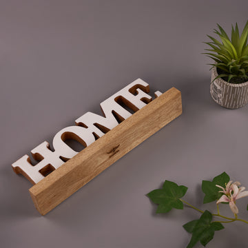 A wooden 'HOME' word sign with a natural finish, displayed on a gray surface with a potted plant and decorative flower nearby.