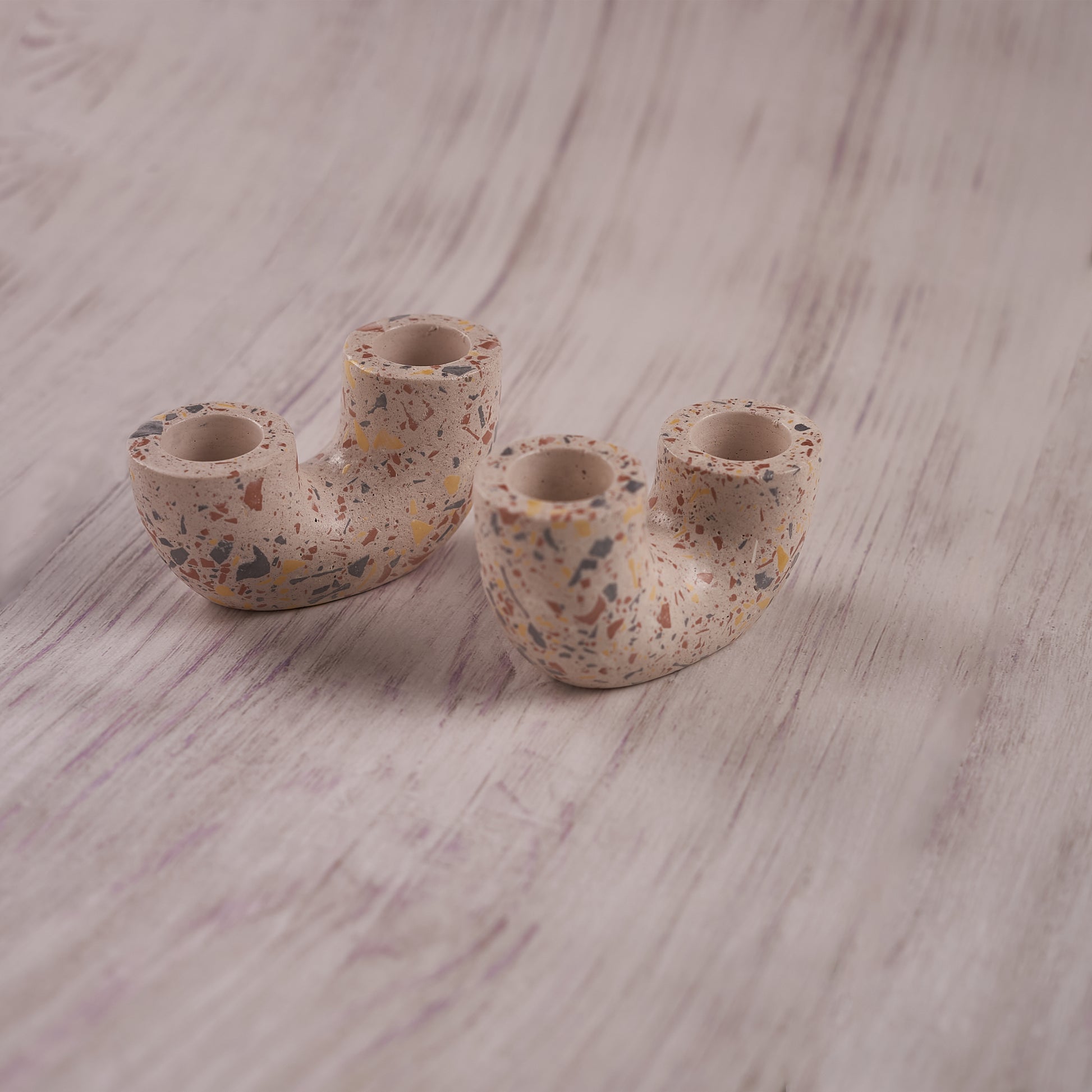 A pair of U-shaped, terrazzo-style Ceramic Curve Candle Holder Set of 2 is displayed on a rustic light wood surface. The speckled design features small, colorful flecks of orange, blue, and yellow, with each holder having two openings for taper candles.