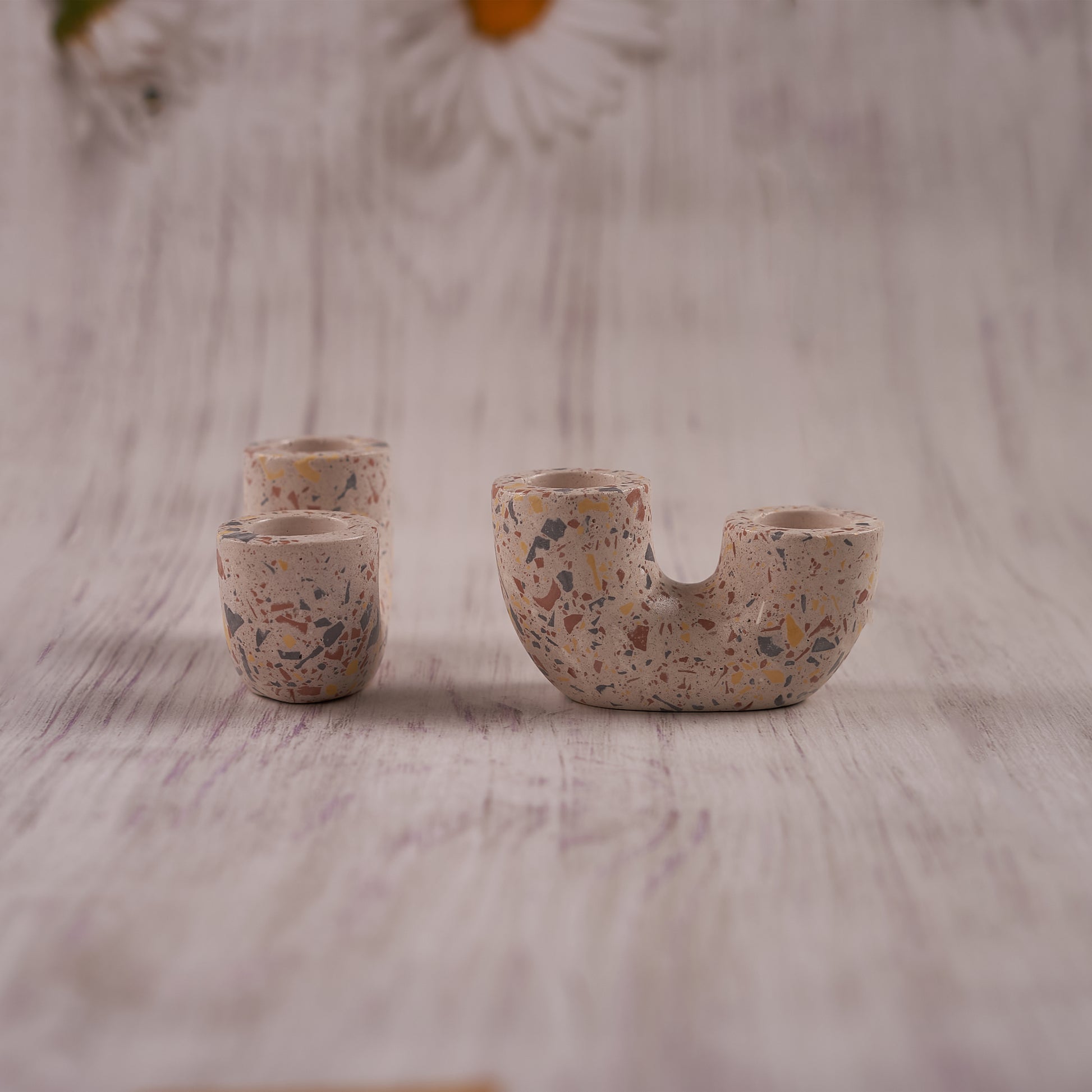 A close-up shot of a unique, speckled Ceramic Curve Candle Holder Set of 2 on a light wooden background. The main U-shaped piece holds two candles, while two smaller cylindrical holders stand nearby, all featuring a colorful terrazzo-like pattern with flecks of red, yellow, and blue.