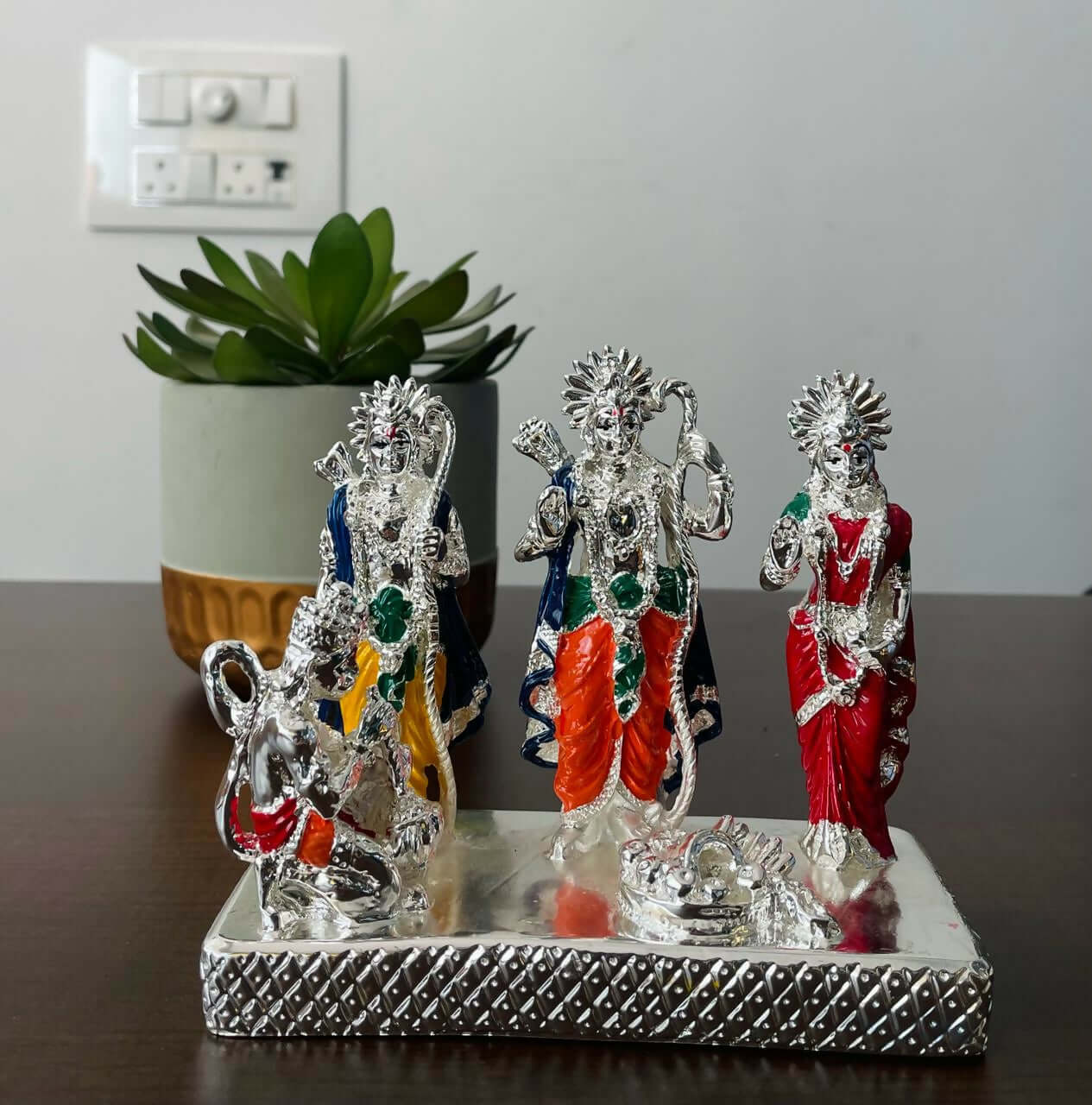 Silver-plated Ram Darbar idol set depicting Lord Rama, Goddess Sita, Lord Lakshmana, and Lord Hanuman, with colored details, placed on a decorative base with a plant in the background.