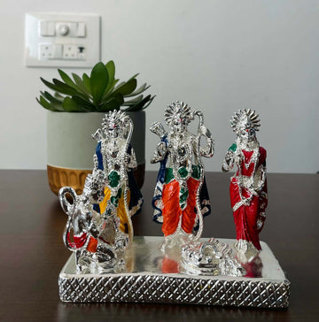 Silver-plated Ram Darbar idol set depicting Lord Rama, Goddess Sita, Lord Lakshmana, and Lord Hanuman, with colored details, placed on a decorative base with a plant in the background.