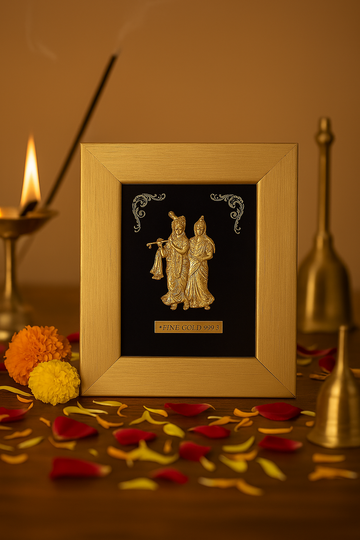 A close-up of a gold-colored photo frame featuring a golden relief of Radha and Krishna, set on a dark wooden surface scattered with red and yellow flower petals and two marigold flowers. In the softly lit background, a lit oil lamp and a burning incense stick are visible, creating a serene, devotional ambiance.