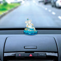 A white and blue Ganesha idol is visible in a car's interior. A blurred view of a road and other cars is visible through the windshield in the background. The idol is on a decorative blue base.