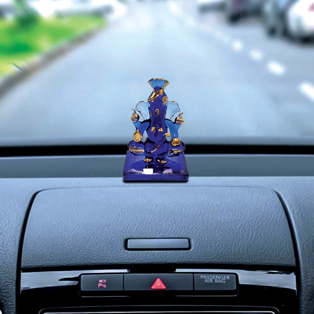A blue and gold Ganesha idol placed on the dashboard of a car, with the road visible through the windshield in the background.
