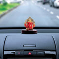 A red and gold Ganesha idol with a detailed gold Kalpavriksha tree as a halo, approximately 3 inches in height, is positioned on a car's dashboard. The background shows a road from the perspective of the driver.