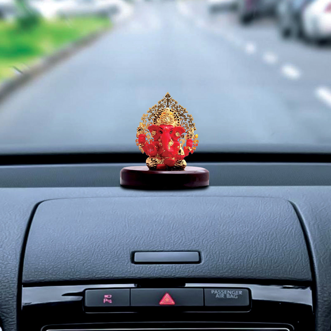 A red and gold Ganesha idol with a detailed gold Kalpavriksha tree as a halo, approximately 3 inches in height, is positioned on a car's dashboard. The background shows a road from the perspective of the driver.