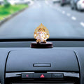 A small Ganesha Idol, approximately 3 inches tall, with a gold-plated Kalpavriksha tree as a halo, sits on a dark wooden base on the dashboard of a car. The idol is white and gold, and the car's interior is gray and black.