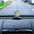 A blue and gold Ganesha idol, approximately 3 inches tall, with a detailed gold Kalpavriksha tree halo is shown on a glossy dark wooden base. The idol is placed on the dashboard of a car, with the road visible through the windshield.