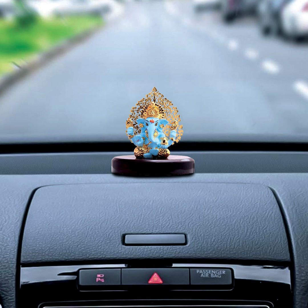 A blue and gold Ganesha idol, approximately 3 inches tall, with a detailed gold Kalpavriksha tree halo is shown on a glossy dark wooden base. The idol is placed on the dashboard of a car, with the road visible through the windshield.