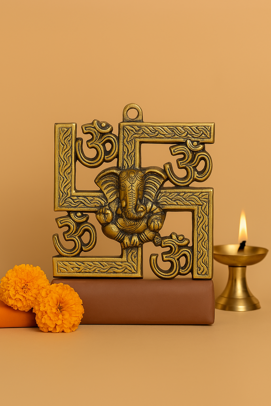 Brass-finished swastika wall hanging with embossed Om symbols and a central Lord Ganesha figure, placed on a brown base with marigold flowers, alongside a lit brass oil lamp against a warm background.