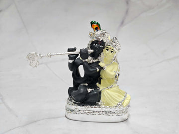 A silver-colored idol depicting Lord Krishna, shown with dark skin and holding a flute, seated beside Radha, who is depicted with lighter, possibly white or silver, skin. The idol rests on a silver-colored base.