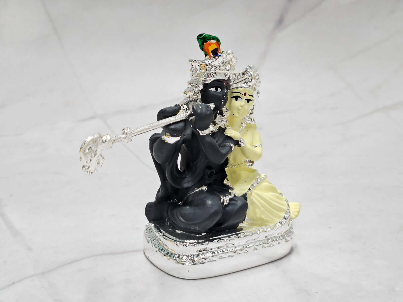 A silver-plated idol of Radha Krishna on a marble surface. Krishna is depicted in dark stone, playing a silver flute, while Radha in light stone sits beside him, adorned with intricate details and a peacock feather on Krishna's crown.