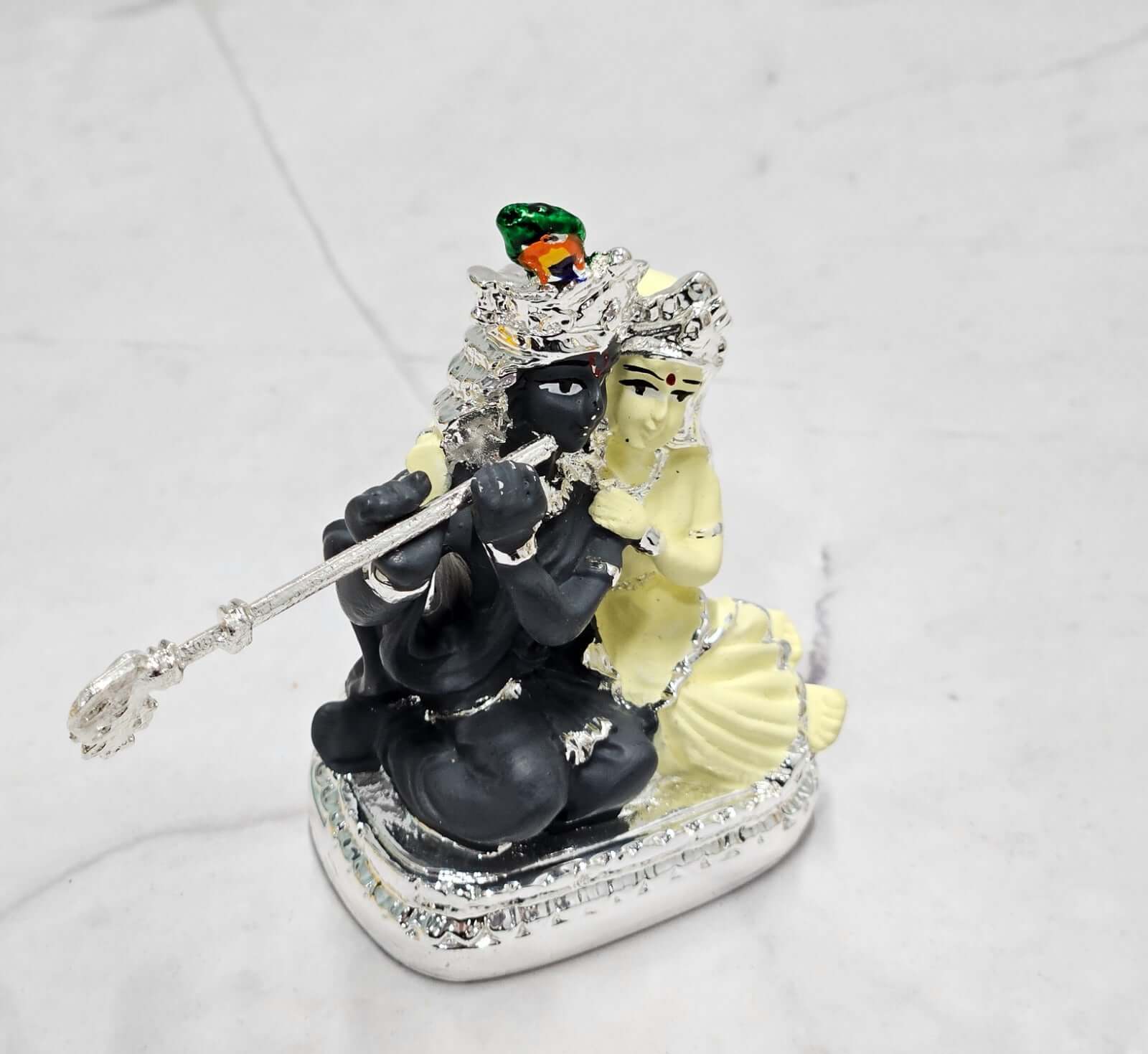 A silver-plated statue depicting the Hindu deities Radha and Krishna. Krishna, with a dark complexion, is playing a silver flute while seated, and Radha, with a light complexion, is gracefully positioned beside him, both adorned with elaborate crowns and jewelry, resting on a decorative base.