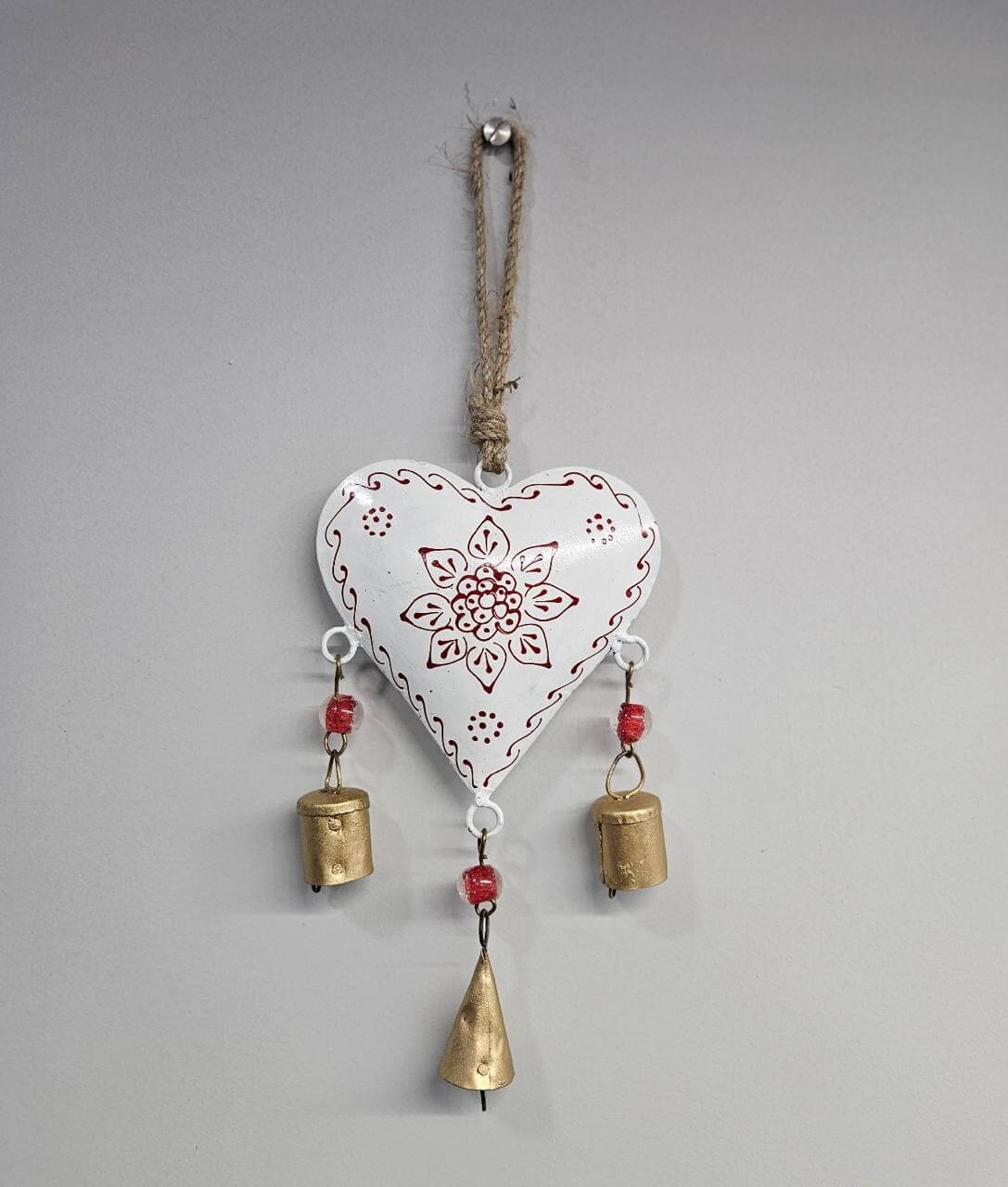 A hand-painted white metal chime with intricate designs, hanging from a jute string, with gold-colored bells attached.