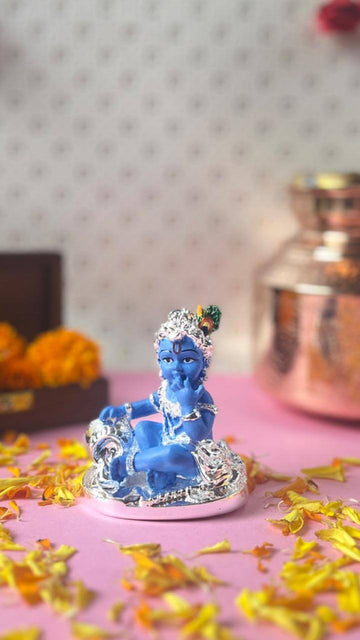 A small blue idol of Bala Krishna, adorned with silver jewelry, sits gracefully on a pink surface surrounded by scattered yellow flower petals. In the blurred background, a warm-toned bronze pot and a box with orange marigold flowers add depth to the scene.