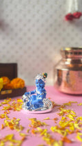 A blue statue of Baby Krishna (Laddu Gopal or Bala Krishna), adorned with a silver crown and jewelry, sits on a silver pedestal covered with scattered yellow and orange flower petals on a pink surface. In the blurred background, a copper pot and a wooden box filled with marigold flowers are visible.