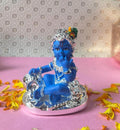 A blue statue of infant Lord Krishna (Bala Krishna) adorned with silver jewelry and a peacock feather, sitting on a decorative base surrounded by yellow and orange flower petals.