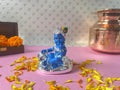 A blue statue of infant Lord Krishna (Bala Krishna) with a makhan matki (butter pot), surrounded by yellow and orange flower petals.
