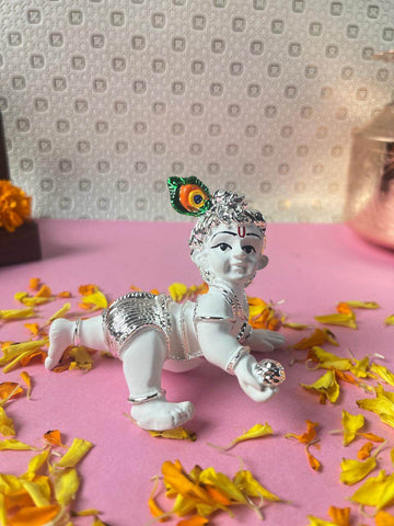 A white idol of crawling Baby Krishna (Laddu Gopal) adorned with silver accents and a peacock feather crown, surrounded by yellow flower petals on a pink surface.