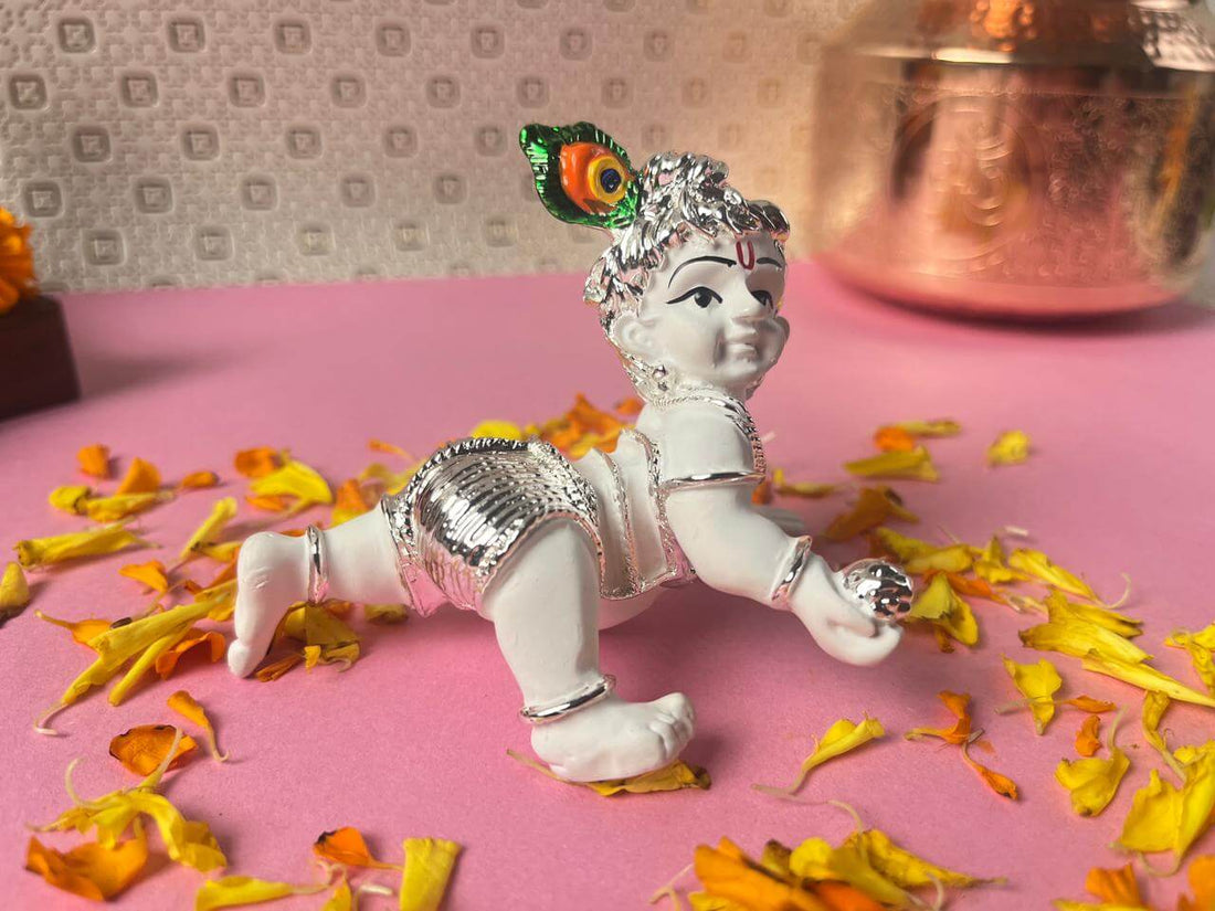 A white idol of crawling Baby Krishna (Laddu Gopal) adorned with silver accents and a peacock feather crown, surrounded by yellow flower petals on a pink surface.