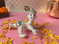 White and silver idol of crawling Bal Krishna holding a silver butter ball, adorned with a colorful peacock feather, surrounded by yellow and orange flower petals on a pink surface.