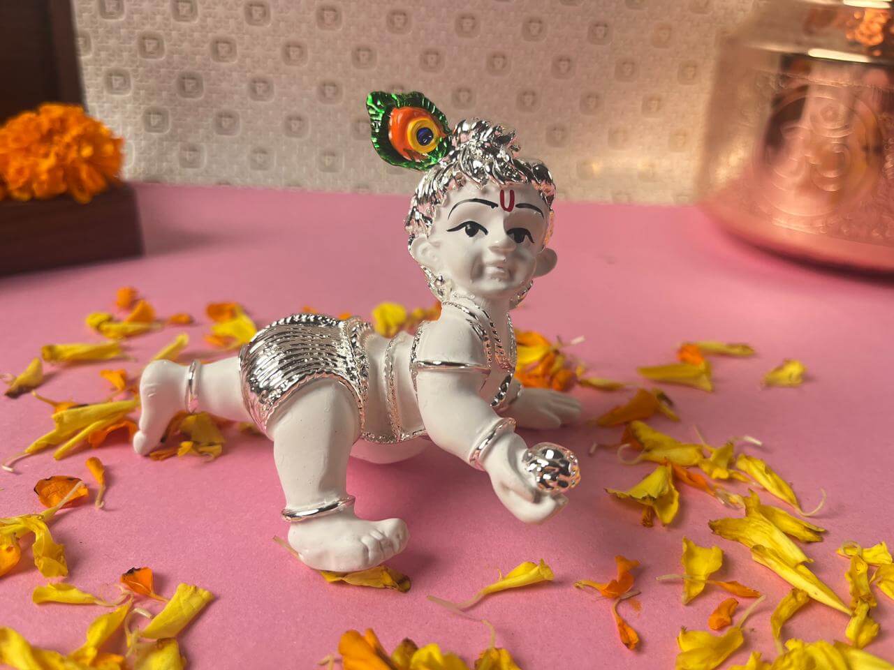 White and silver idol of crawling Bal Krishna holding a silver butter ball, adorned with a colorful peacock feather, surrounded by yellow and orange flower petals on a pink surface.