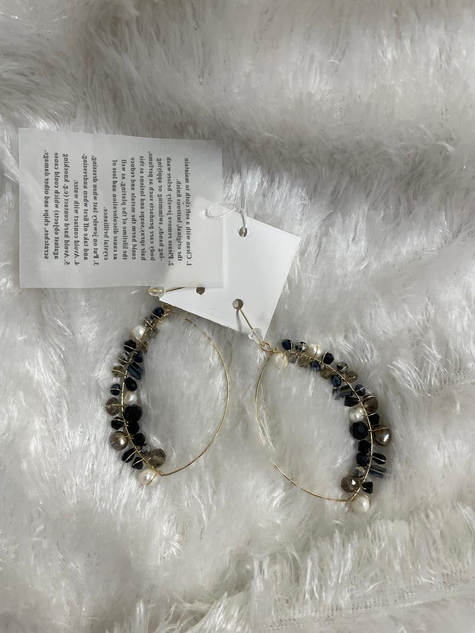 A pair of large, gold-toned hoop earrings, adorned with a cluster of various beads, are displayed on a soft, light-colored background. The beads include small white pearls and faceted Crystal Bead Wrapped Earrings in Black & Grey, in different shapes and sizes, which are wrapped along one side of each hoop.