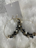 A pair of large, gold-toned hoop earrings are displayed on a soft, light-colored, fluffy surface. The lower half of each hoop is embellished with a cluster of various faceted and smooth beads, including white pearls and Crystal Bead Wrapped Earrings in Black & Grey, creating a striking, asymmetrical design.
