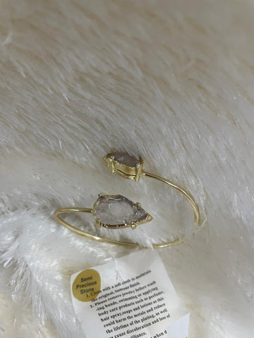 A rose quartz arrowhead rough stone cuff bracelet in a light gold metal finish, presented on a white fuzzy surface with a care instruction tag.