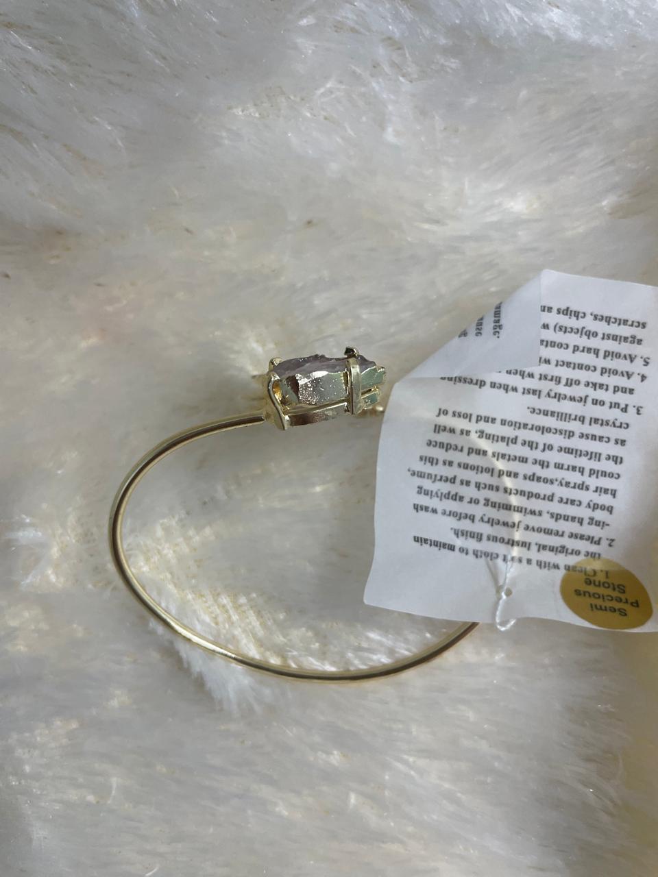 A rose quartz arrowhead rough stone cuff bracelet with a light gold metal finish, displayed on a soft white textured surface alongside a care instruction tag.
