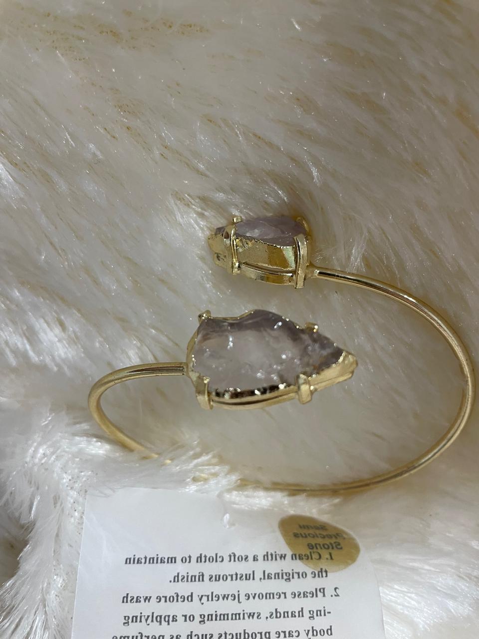 A cuff bracelet featuring two rough rose quartz arrowhead stones set in a light gold metal finish, displayed on a fluffy white fabric surface.