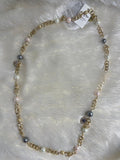 A Brass Long Chain and Swarovski pearl Necklace is shown lying on a fluffy white surface. The long gold-toned chain is interspersed with various sizes and colors of pearls, including white, light pink, and grey.