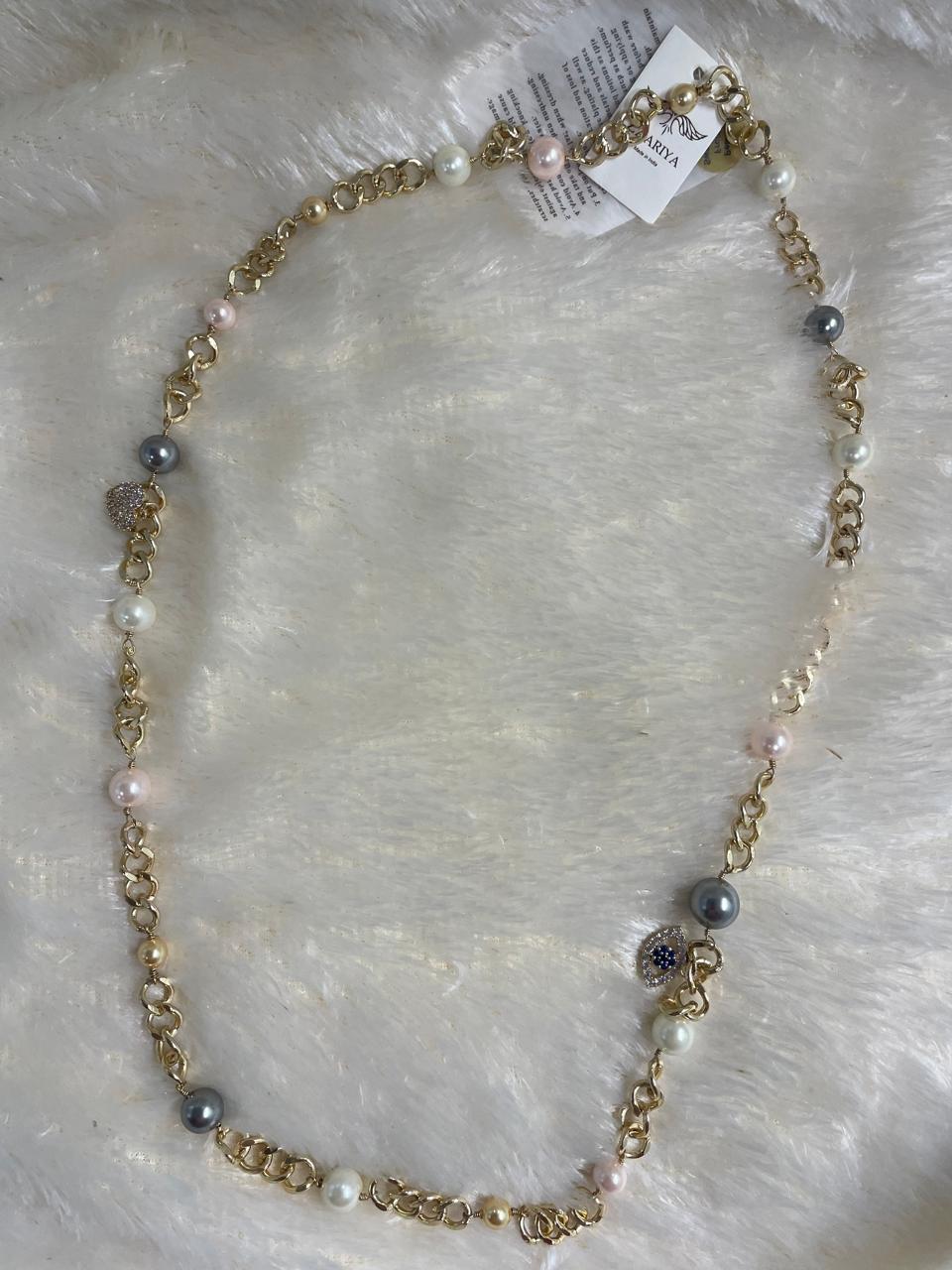 A Brass Long Chain and Swarovski pearl Necklace is shown lying on a fluffy white surface. The long gold-toned chain is interspersed with various sizes and colors of pearls, including white, light pink, and grey.