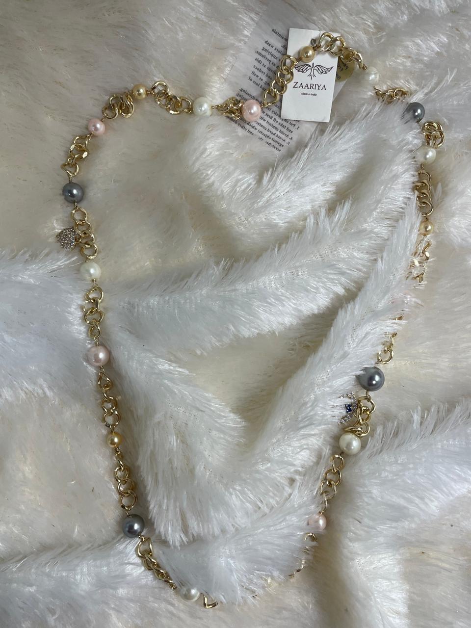 A brass long chain necklace with Swarovski pearl beads on a white furry surface.