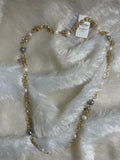 A Brass Long Chain and Swarovski pearl Necklace is shown lying on a fluffy white surface. The long gold-toned chain is interspersed with various sizes and colors of pearls, including white, light pink, and grey.