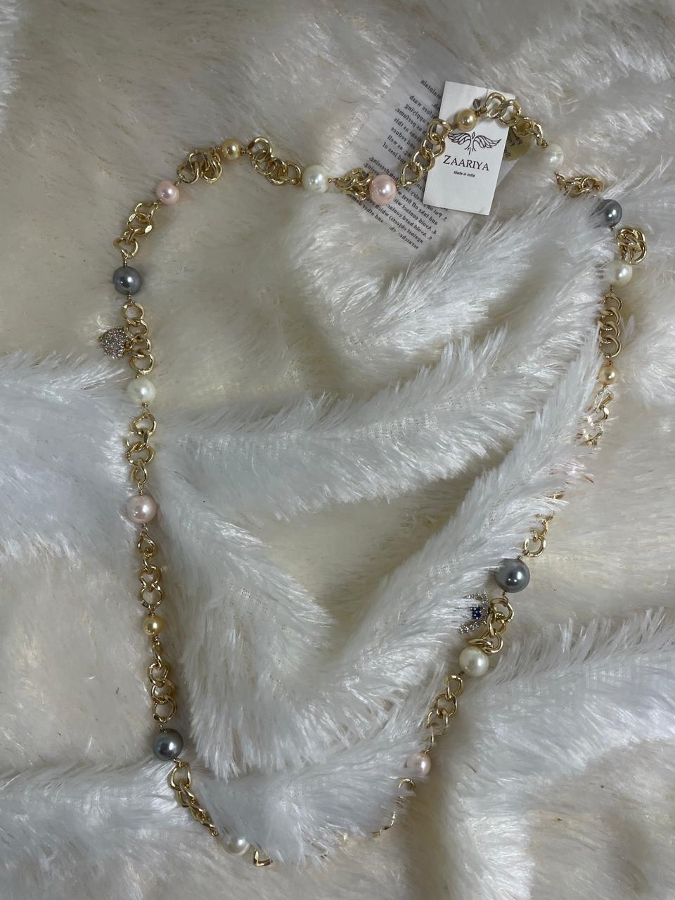 A Brass Long Chain and Swarovski pearl Necklace is shown lying on a fluffy white surface. The long gold-toned chain is interspersed with various sizes and colors of pearls, including white, light pink, and grey.