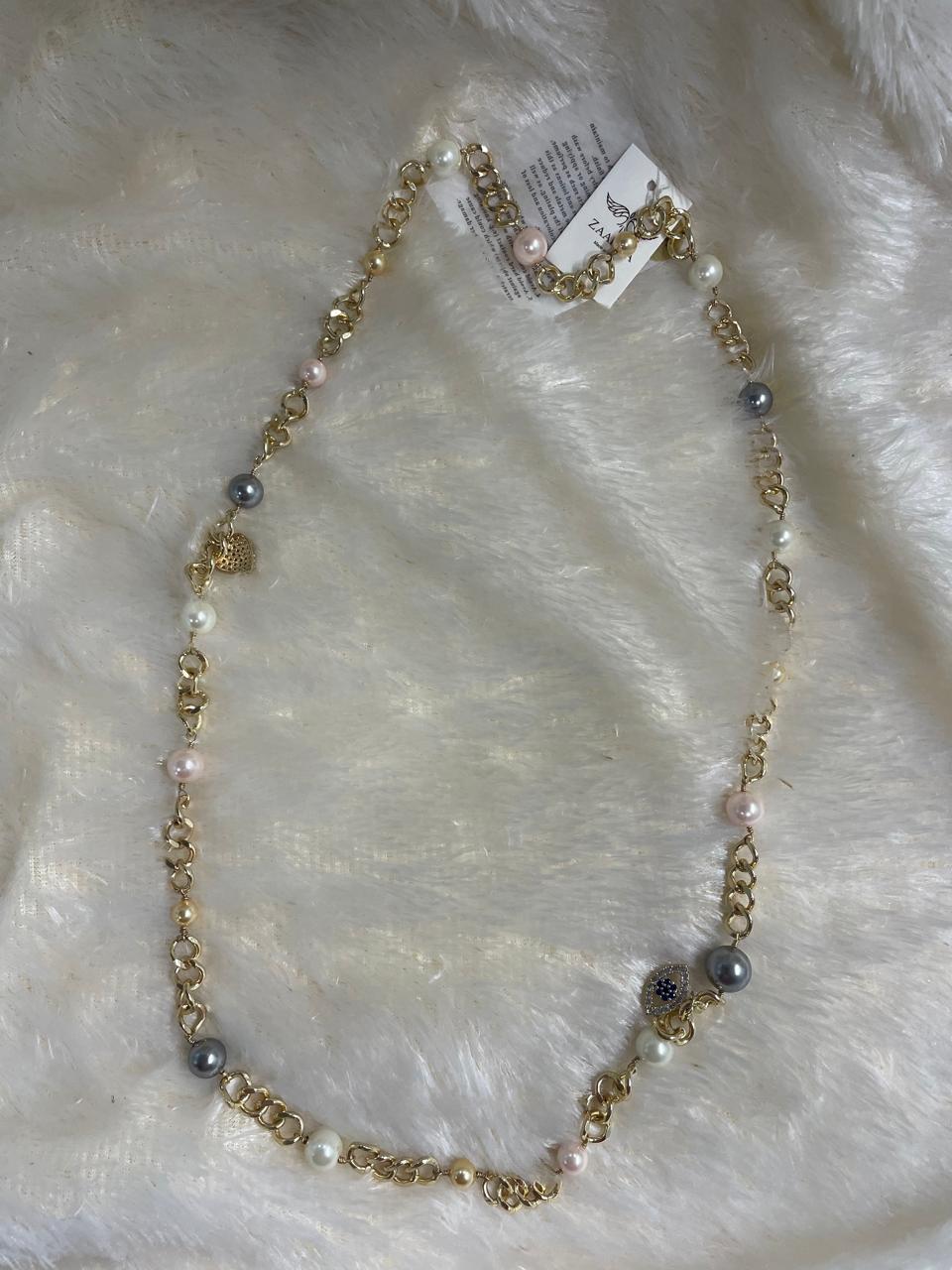A brass long chain necklace with Swarovski pearl beads on a white furry surface.