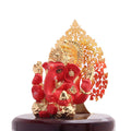 A red and gold Ganesha idol, seated on a dark base, with a golden, intricately designed tree-like structure forming a backdrop behind the deity.