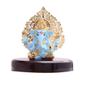 A blue and gold Lord Ganesha idol with a detailed, tree-like golden arch behind him, sitting on a dark wooden base, typically used for car dashboards or home decor.