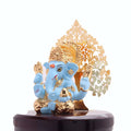 A close-up of a vibrant blue Lord Ganesha idol with golden adornments, seated on a dark base, in front of an intricate golden tree-shaped backdrop, likely a decorative piece for a car dashboard or home.