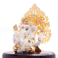 White and gold Ganesha idol sitting on a dark wooden base with a golden, intricate backdrop.