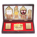 A decorative wooden box with a glass top displays gold-plated idols of Lord Jagannath, Lord Balabhadra, and Goddess Subhadra, alongside a golden Charan Paduka and inscribed plaques with Sanskrit text on a red velvet lining