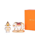 A close-up of a decorative display featuring a gold and white Kamdhenu cow with calf idol, an ornate golden and silver vessel, and a partially visible orange box with the 'WEMY' logo.