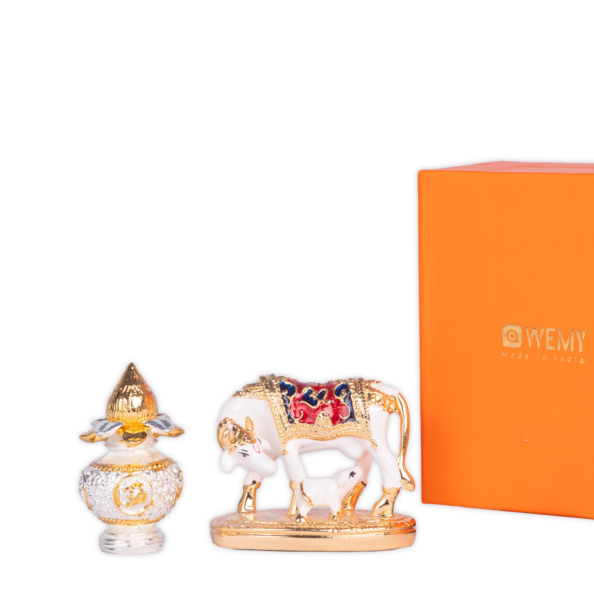 A close-up of a decorative display featuring a gold and white Kamdhenu cow with calf idol, an ornate golden and silver vessel, and a partially visible orange box with the 'WEMY' logo.
