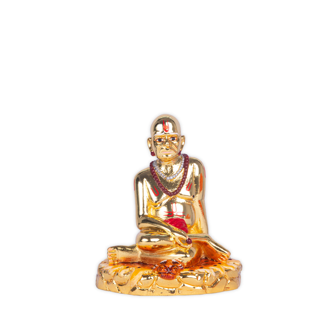 A close-up of a golden Swami Samarth idol seated in a meditative pose, adorned with a red tilak and garland, surrounded by traditional offerings like marigold petals, red rose petals, and ladoos, against a warm yellow background.