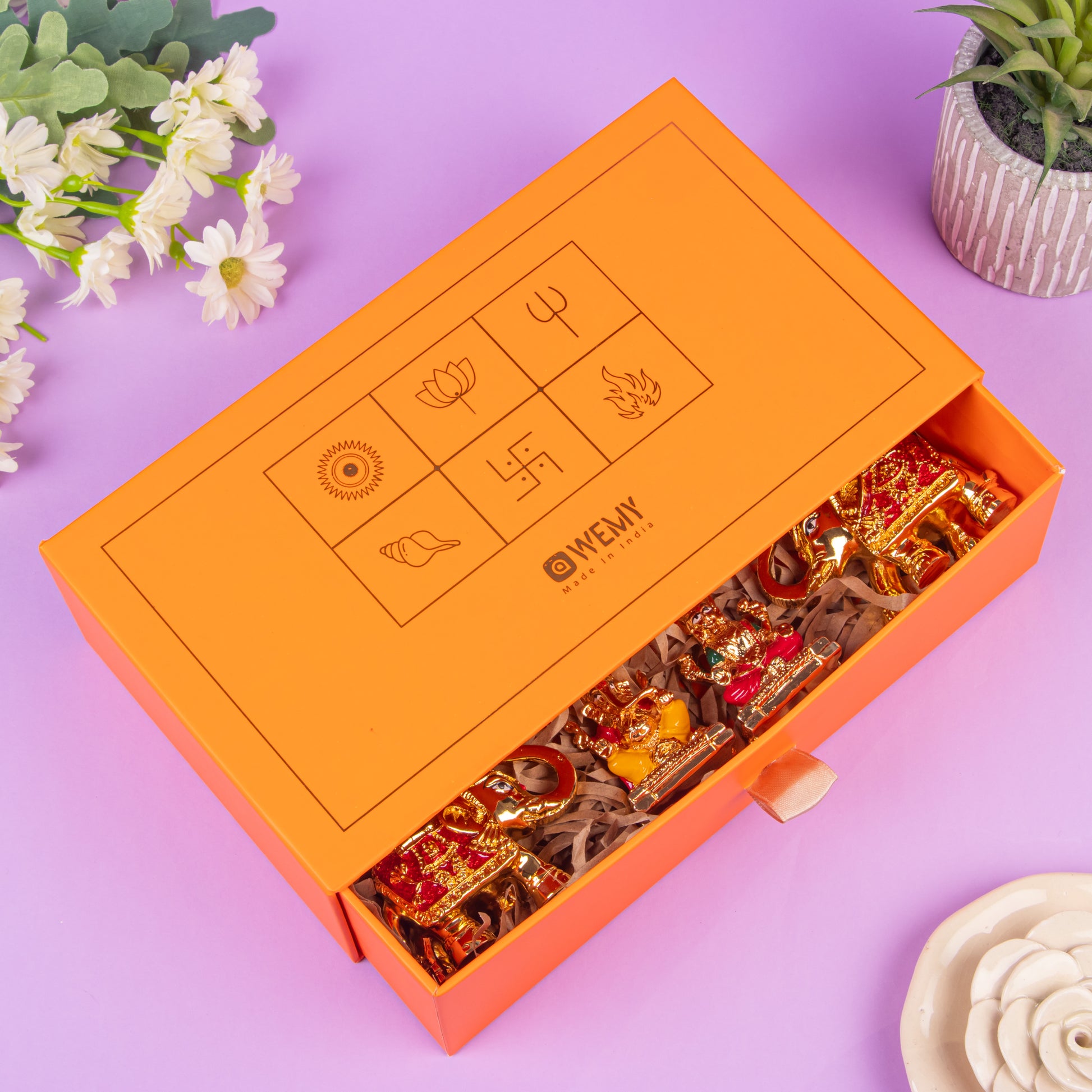 Partially opened orange gift box with traditional Indian symbols on the lid, revealing gold-plated decorative figurines inside, placed on shredded paper, surrounded by flowers and a small potted plant on a purple background.