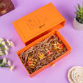 Elegant orange gift box with gold-plated decorative figurines of two elephants and a deity, presented on shredded paper, surrounded by flowers and plants on a purple background.